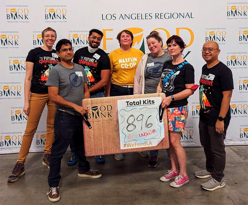 Seven MaxFun worker-owners standing at the Los Angeles Regional Food Bank, holding a sign that reads "Total Kits: 896. Wow! #WeFeedLA"