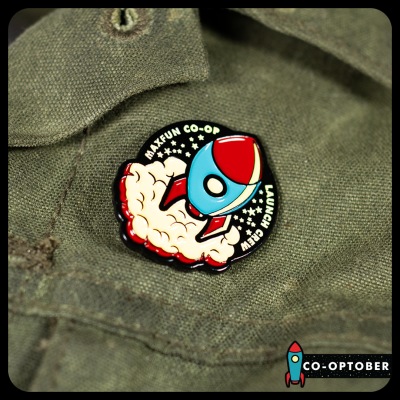 A photo of our pin which depicts a rocket in three quarter view blasting off with a cloud of smoke behind it. Text: MaxFun Launch Crew